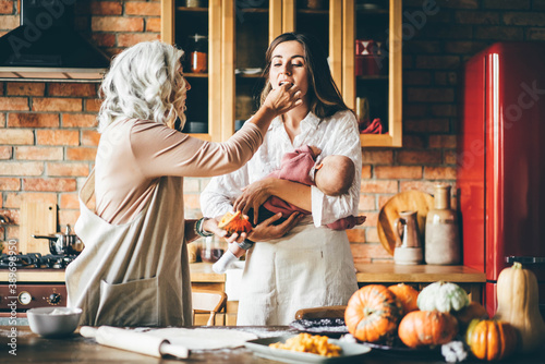 Brunette with long loose flowing hair holds baby and senior granddaughter preparing dinner in kitchen.