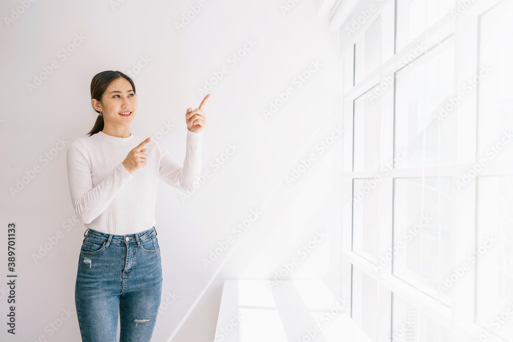 Good looking Asian female wearing a sweater and jeans point her fingers in the white wall and window, standing near the window with natural light. People in the indoor background with copy-space. Good looking Asian female wearing a sweater and jeans point her fingers in the white wall and window, standing near the window with natural light. People in the indoor background with copy-space.