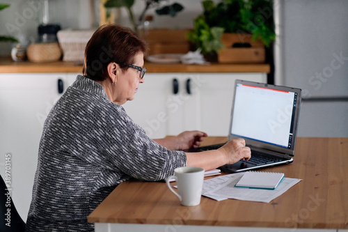 Senior woman using laptop for websurfing in her kitchen. The concept of senior employment, social security. Mature lady sitting at work typing a notebook computer in an home office.