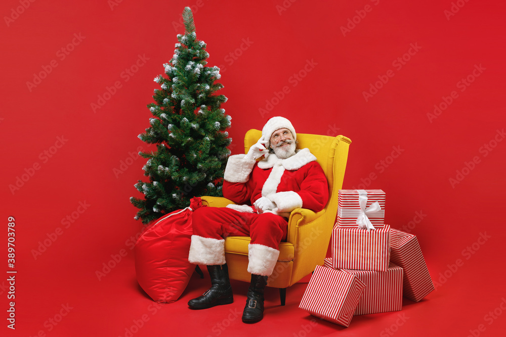 Pensive Santa Claus man in Christmas hat suit coat sit in armchair with fir tree presents gifts put hand on head isolated on red background studio. Happy New Year celebration merry holiday concept.