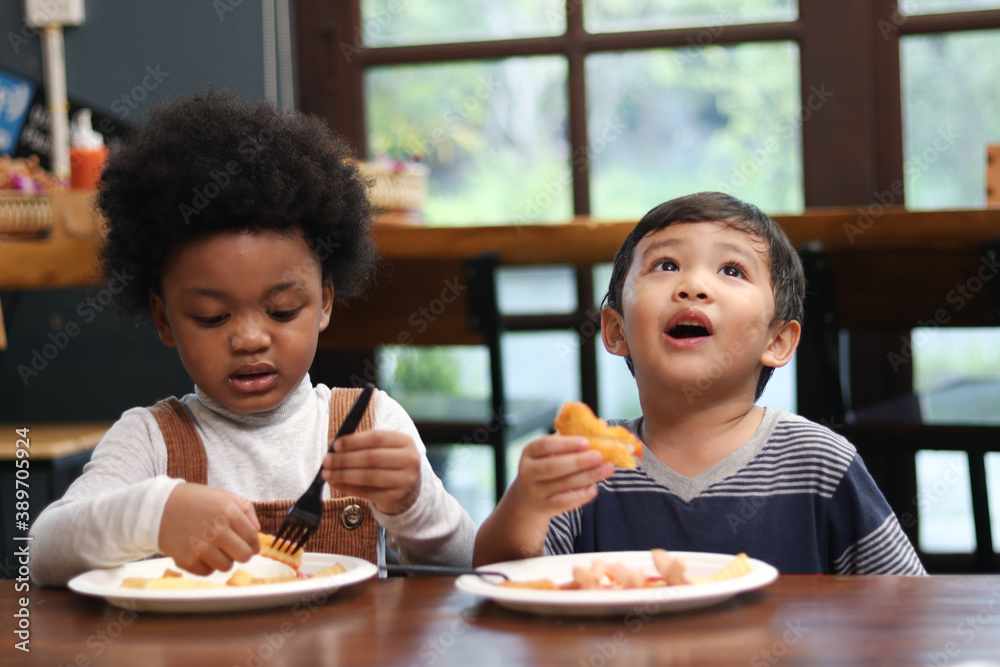 Cute African American boy with curly and adorable Asian kid eating meal at the table indoor, happy children having food in a restaurant, enjoy their meal together Cute African American boy with curly and adorable Asian kid eating meal at the table indoor, happy children having food in a restaurant, enjoy their meal together