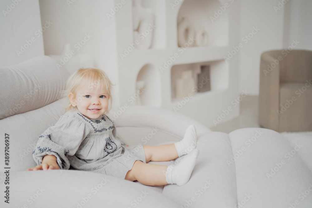 portrait of a little daughter, a blonde girl in a stylish beige dress, playing fun games at home on the sofa and smiling. bright and stylish interior of the apartment portrait of a little daughter, a blonde girl in a stylish beige dress, playing fun games at home on the sofa and smiling. bright and stylish interior of the apartment
