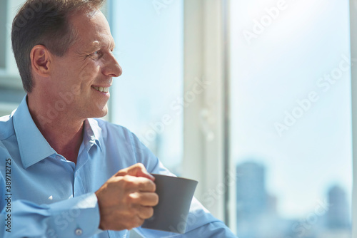 Close up shot of a happy mature businessman drinking fresh morning coffee in the office, looking in window and enjoying city view
