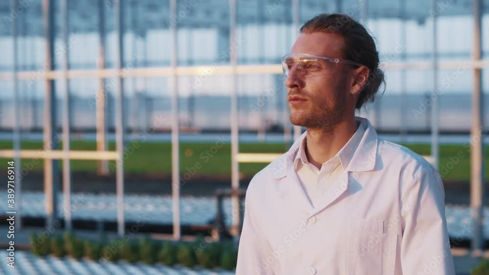Young greenhouse expert bearded caucasian worker in eyeglasses walking in the farm construction garden examining plants. Professional worker. Agriculture concept. Young greenhouse expert bearded caucasian worker in eyeglasses walking in the farm construction garden examining plants. Professional worker. Agriculture concept.