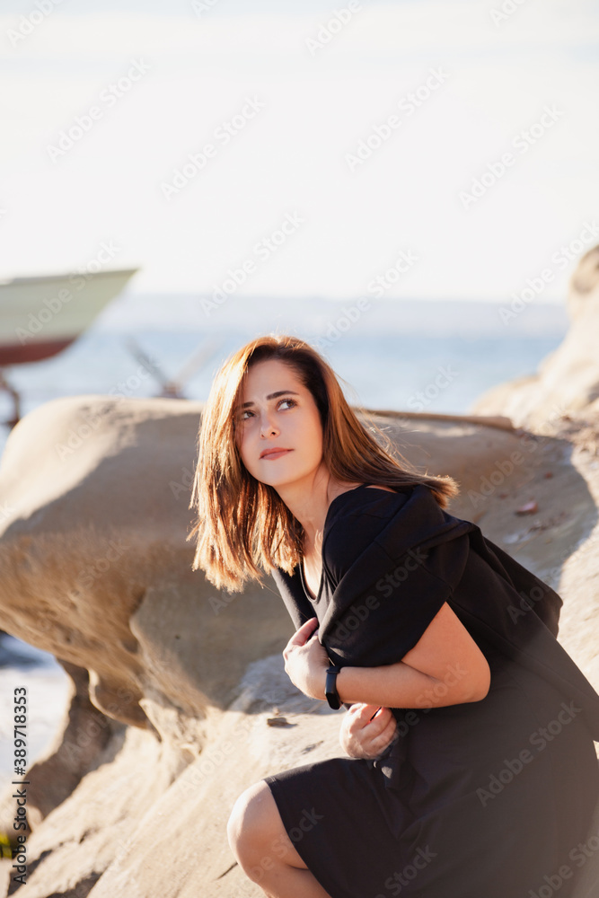 Beautiful dreamy young woman in black clothes sitting on the beach rocks. Sea and boats on the background.