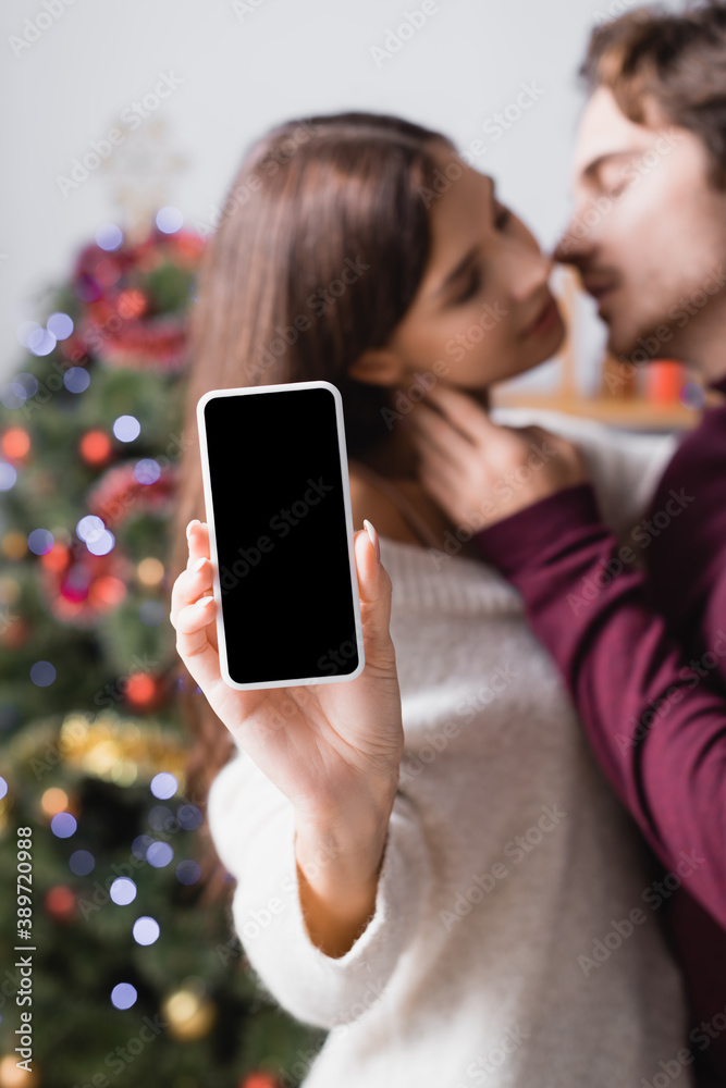 woman holding smartphone with blank screen near boyfriend and decorated christmas tree on blurred background