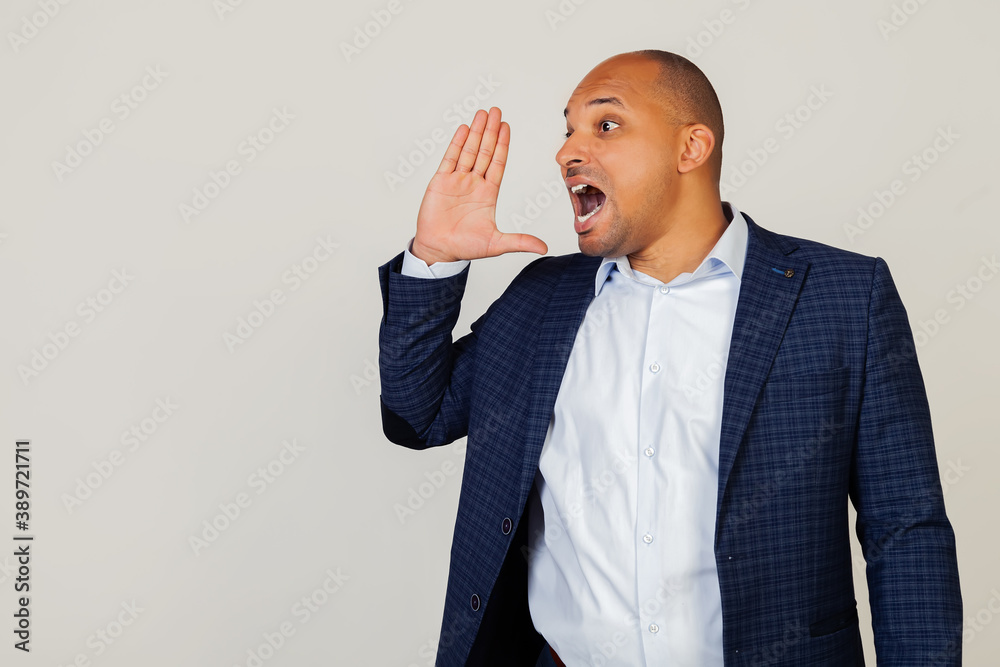 Portrait of a successful young African American businessman guy yelling and shouting loudly to the side with hand over his mouth. Communication concept. Standing on a gray background Portrait of a successful young African American businessman guy yelling and shouting loudly to the side with hand over his mouth. Communication concept. Standing on a gray background