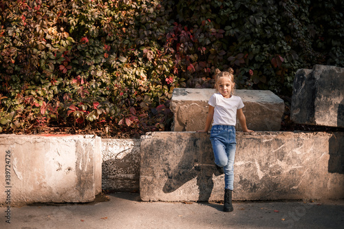 Street photo of a little beautiful girl in jeans and a white t-shirt on a background of concrete slabs and autumn leaves © Дмитрий Ткачук