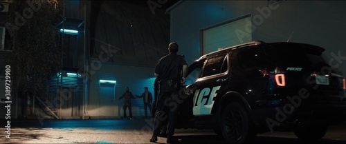 WIDE Police officer exiting a car and approaching two multi-ethnic males suspects near industrial buildings at night. Shot with anamorphic lens © supamotion