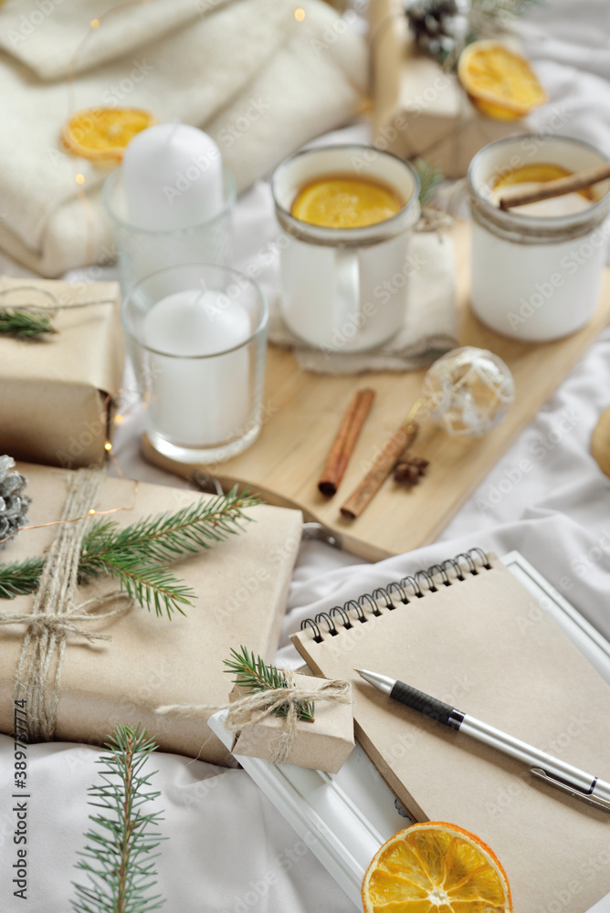 Cosy home flat lay composition with present boxes, blank copybook, a pen, tea and cones on a bed. Holiday shopping list, wish list or New Year Goals concept, presents checklist. Selective focus Cosy home flat lay composition with present boxes, blank copybook, a pen, tea and cones on a bed. Holiday shopping list, wish list or New Year Goals concept, presents checklist. Selective focus