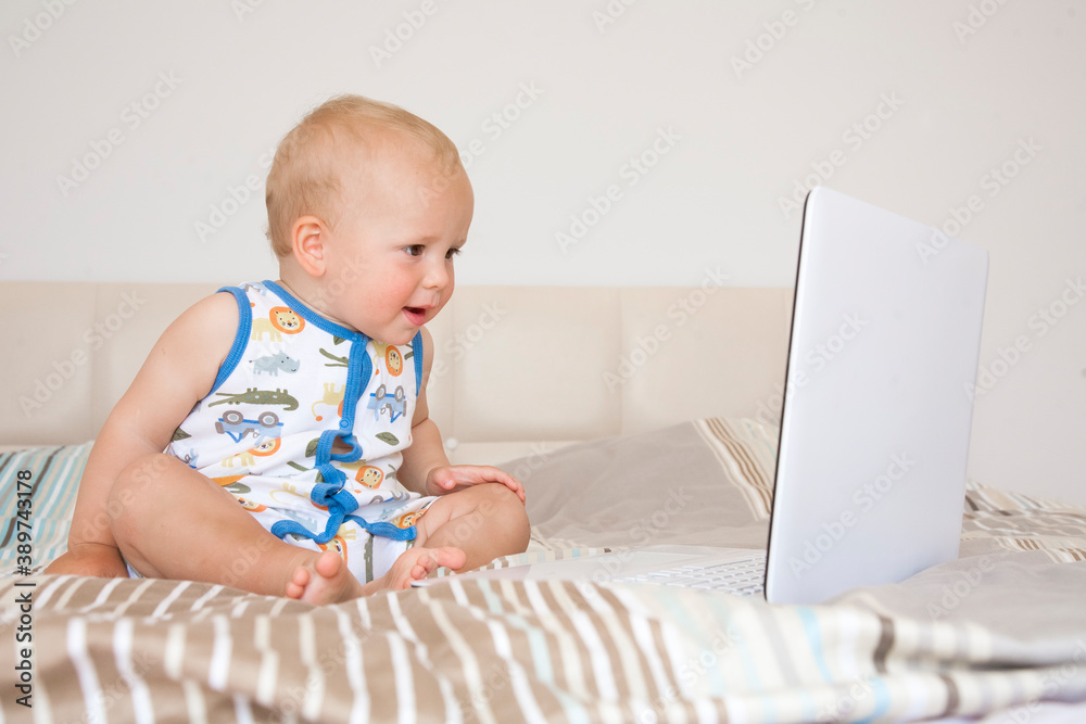 Adorable cute child boy sits on a bed in front of an open computer laptop and emotionally looks at the screen.
Adorable cute child boy sits on a bed in front of an open computer laptop and emotionally looks at the screen.