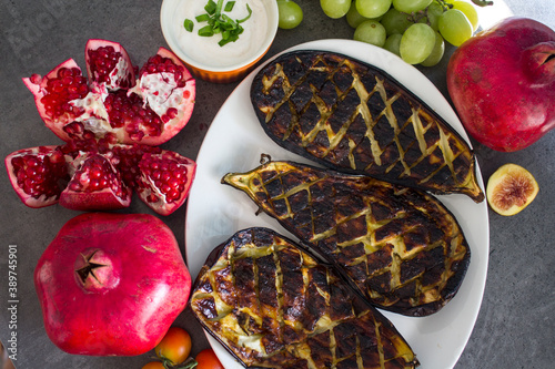 Roasted eggplants on white plate, pomegranates and grapes on a table. Mediterranean style food. 