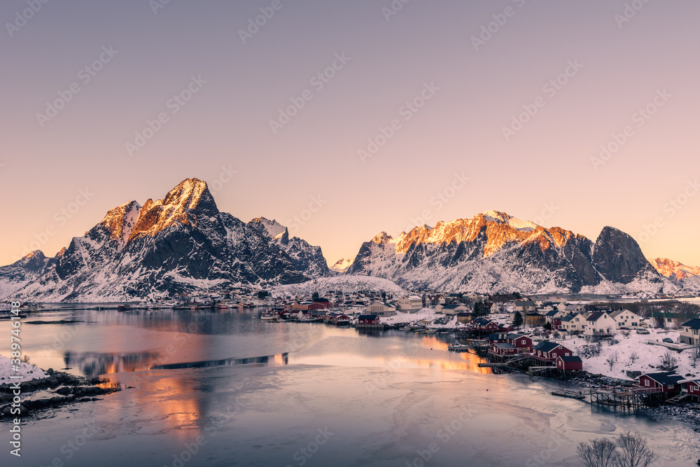 The small fishing village Reine on the Lofoten islands in Norway in winter with steep snowcapped mountains and frozen lake during sunset