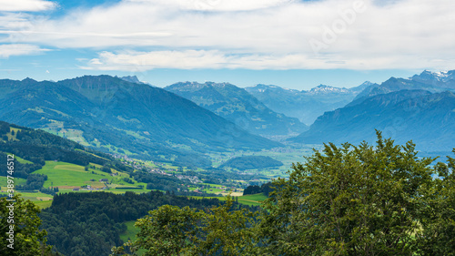 Paysage de montagne avec une vallée verdoyante, des arbres et de la forêt et des montagnes avec des cimes enneigées à l'arrière plan dans un ciel nuageux.