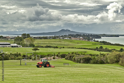 AUCKLAND, NEW ZEALAND - Mar 28, 2019: Lawn mower red tractor working in Macleans park with Hauraki Gulf islands in background photo