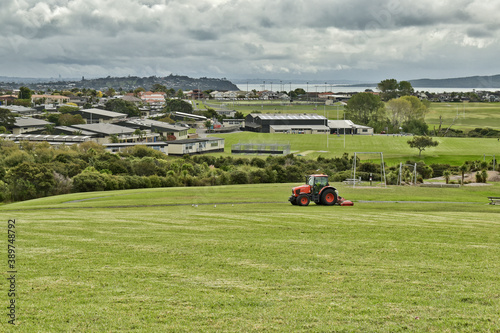 AUCKLAND, NEW ZEALAND - Mar 28, 2019: Lawn mower red tractor working in Macleans park with Hauraki Gulf islands in background photo