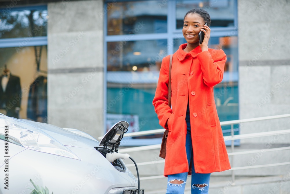 Fototapeta premium Using smartphone while waiting. African American girl on the electric cars charge station at daytime. Brand new vehicle.