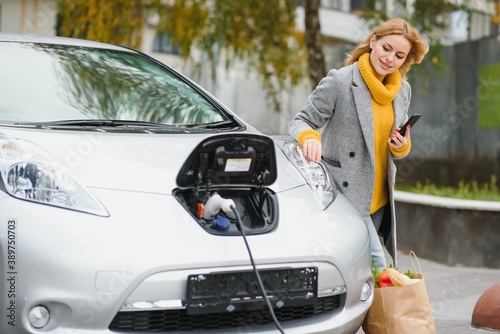 Using smartphone while waiting. Woman on the electric cars charge station at daytime. Brand new vehicle.