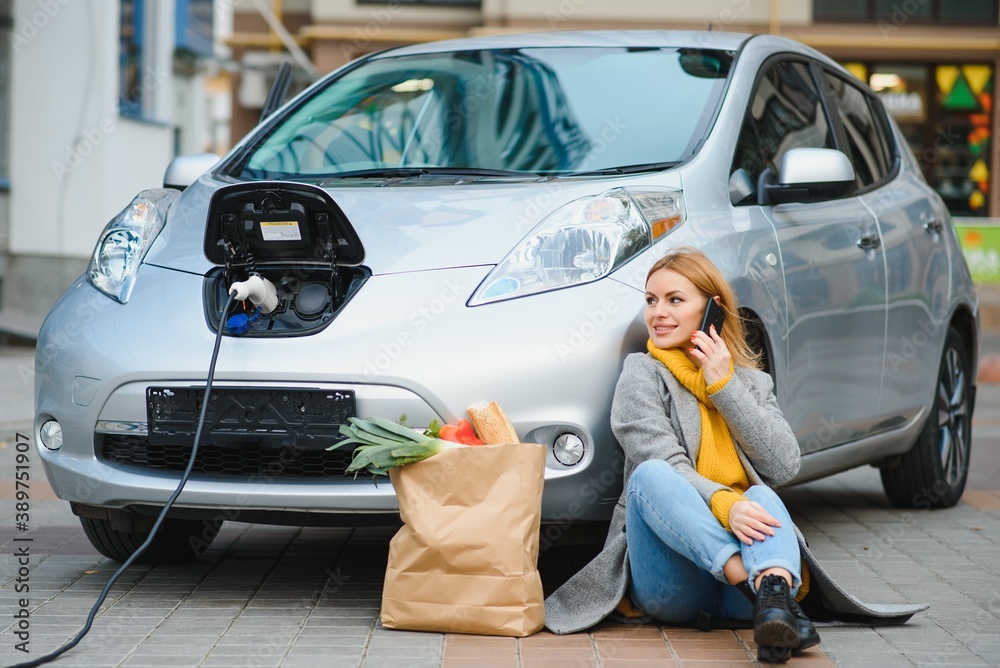 Naklejka premium Having conversation by using phone. Woman on the electric cars charge station at daytime. Brand new vehicle.