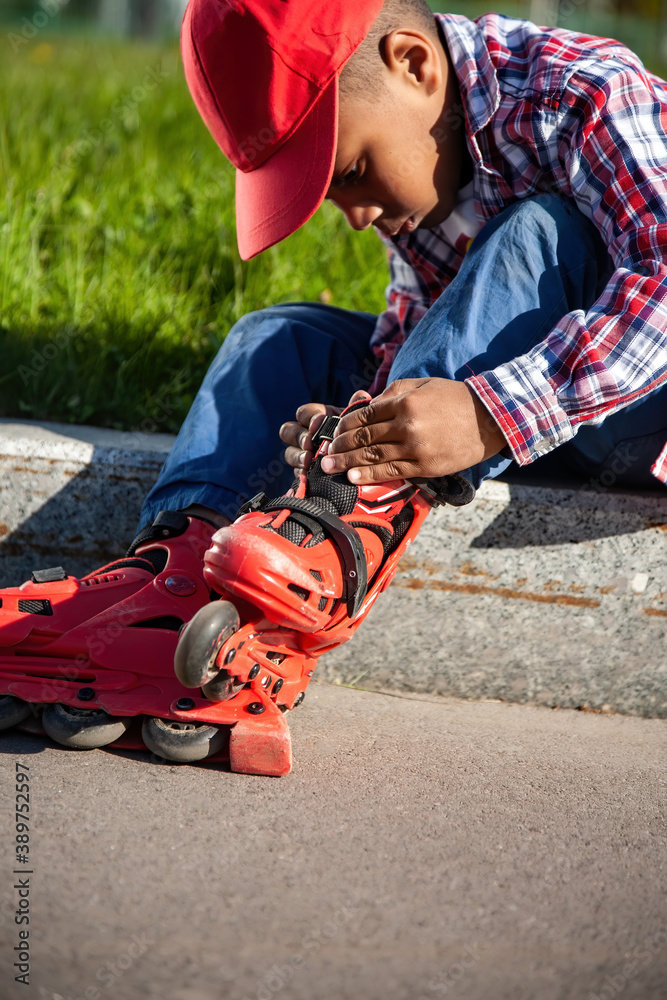 Fototapeta premium First grade little black boy (african american) puts on, adjusts and setting laces on red roller skates before ride. Vertical image.