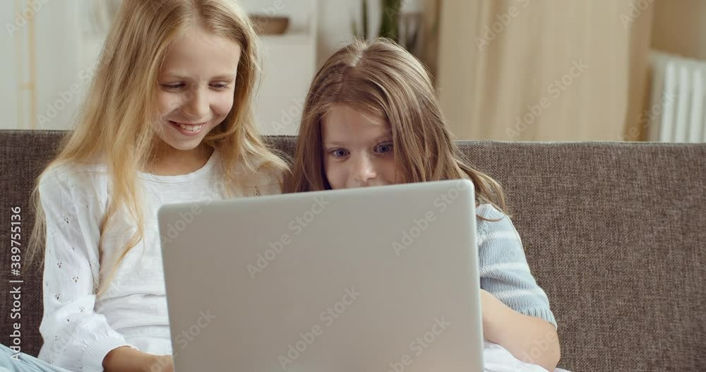 Two happy cute sisters children kid using laptop computer for remote distant education having fun playing or studying together at home sitting at sofa couch. Small caucasian girls having fun in net Two happy cute sisters children kid using laptop computer for remote distant education having fun playing or studying together at home sitting at sofa couch. Small caucasian girls having fun in net