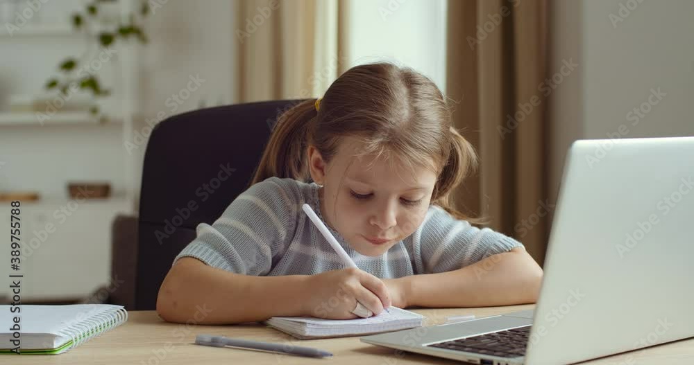 Portrait of cute studious young girl caucasian child using laptop computer speaking with teacher online and writing on notebook, learning and doing homework sitting at home table, coronavirus epidemic Portrait of cute studious young girl caucasian child using laptop computer speaking with teacher online and writing on notebook, learning and doing homework sitting at home table, coronavirus epidemic