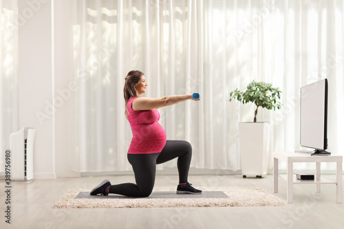 Pregnant woman kneeling and exercising with dumbbells in front of tv