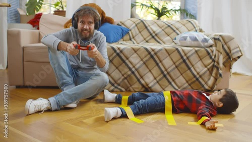 Father in headphones playing video game with console and disobedient son tied with tape on floor photo
