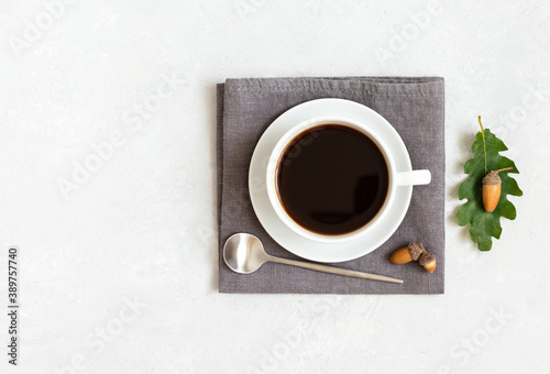 A cup of black coffee on a gray napkin with a teaspoon, acorns and oak leaves. Acorn coffee without caffeine on a white background. Copy space, top view.