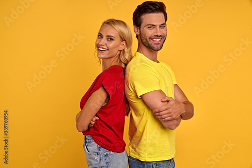 Side view of smiling young couple in red and yellow t-shirts isolated on yellow background. People lifestyle concept. Mock up copy space. Standing back to back holding hands crossed looking camera.