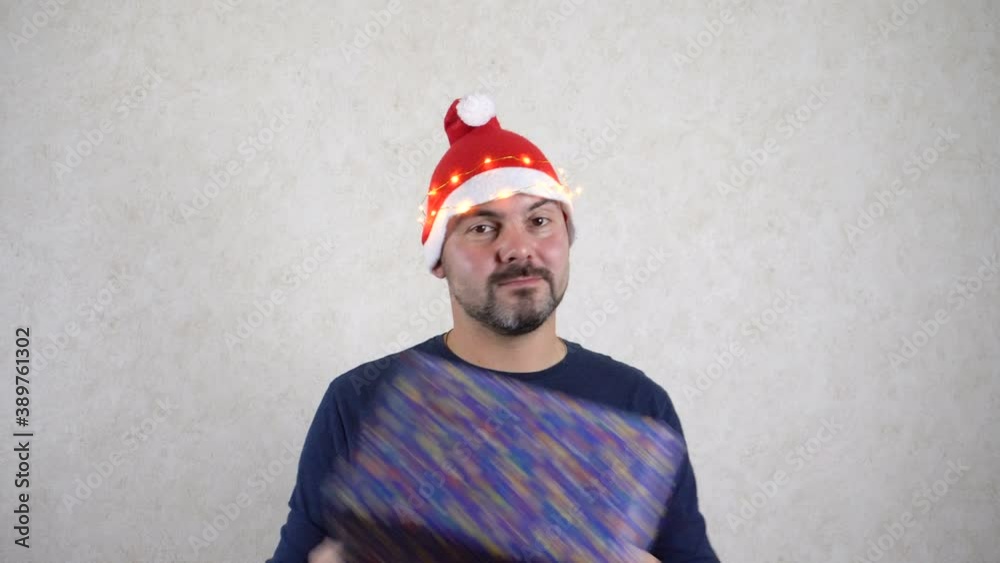 A bearded man in a New Year's hat is dancing with a gift. Good mood. Christmas and New Years holiday. Light background. A bearded man in a New Year's hat is dancing with a gift. Good mood. Christmas and New Years holiday. Light background.