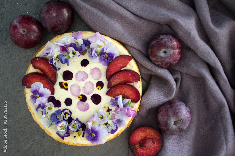 Fototapeta premium Cheesecake decorated with red plums and edible flowers. Fabric texture background. Top view photo of homemade dessert. 