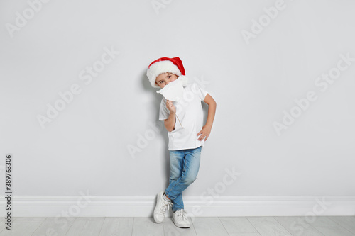 Cute little boy with Santa hat and white beard prop near white wall. Christmas celebration