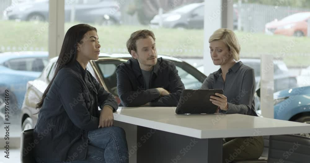 Saleswoman advising couple on car purchase in vehicle dealership showroom using digital tablet Saleswoman advising couple on car purchase in vehicle dealership showroom using digital tablet