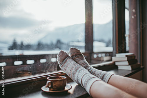 Feet in woollen socks by the Alps mountains view. Woman relaxes by mountain view with a cup of hot drink. Close up on feet. Winter and Christmas holidays concept.