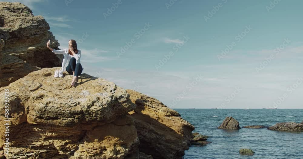 Camera moves around beautiful caucasian woman sitting on sea rock, holding smartphone and shooting video on camera. Camera moves around beautiful caucasian woman sitting on sea rock, holding smartphone and shooting video on camera.