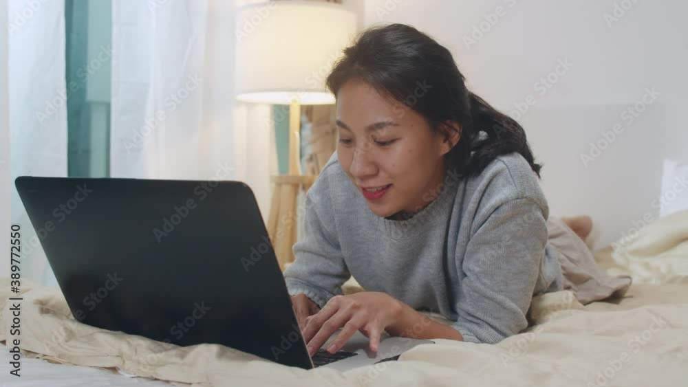 Portrait of freelance Asia woman casual wear using laptop on bed in bedroom at house night. Working from home, remotely work, self isolation, social distancing, quarantine for corona virus prevention.