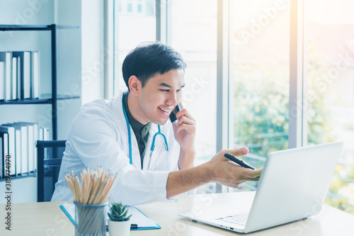 Portrait handsome young medical Asian doctor smiling has stethoscope talking smartphone provide patient consultation check symptom disease on laptop notebook on desk office clinic. Healthcare concept.