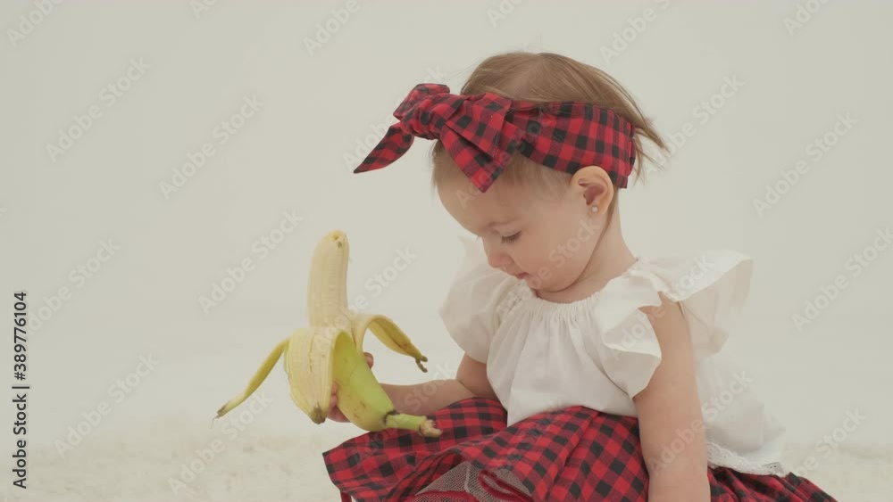 Little girl in plaid headband and skirt is holding big peeled banana. Close up slow motion