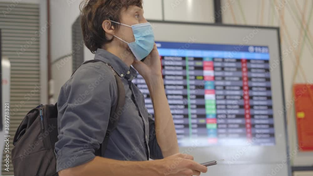 A young man wearing a medical face mask in an airport stands in a hall in front of a flight schedule monitor and talks on a smartphone. The concept of the New normal of people's lifestyle. Air travel
