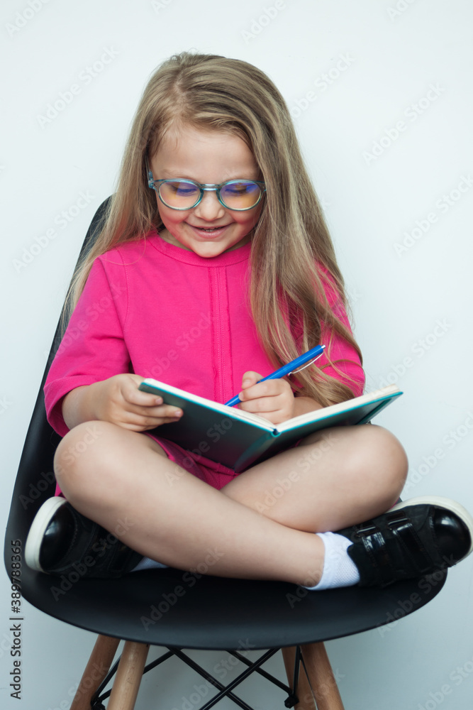 blonde girl with glasses is writing something in a book while posing in an armchair on a white studio wall blonde girl with glasses is writing something in a book while posing in an armchair on a white studio wall