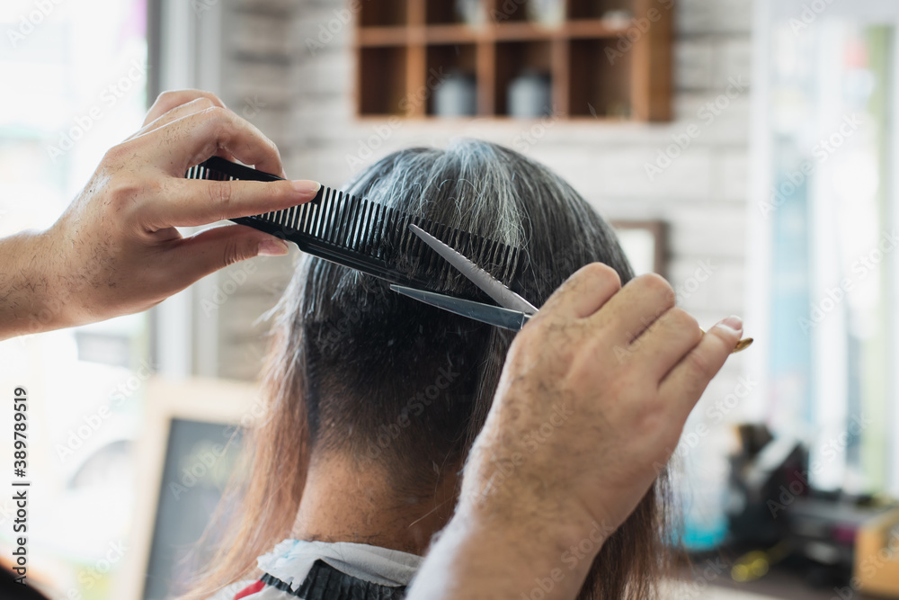 Fototapeta premium Asian young man being cut from long hair to short hair with scissor by professional barber in barbershop.