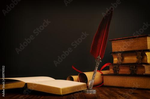 фотографія Old feather quill ink pen with inkwell and old books over wooden desk in front of black wall background
