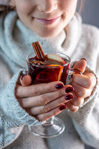 A woman in a white warm sweater is holding a transparent glass cup of mulled wine in her hand. Concept of a cozy atmosphere and a traditional winter drink