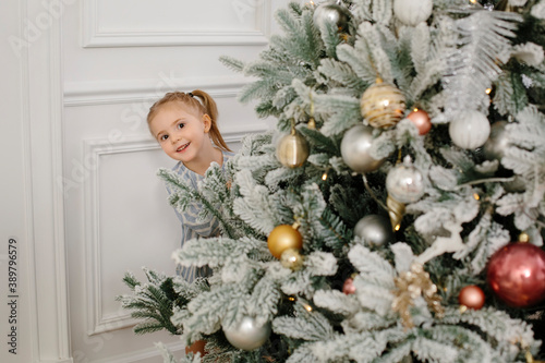 A little cute girl looks out from behind a beautiful Christmas tree. Happy three year old child. Christmas and new year concept