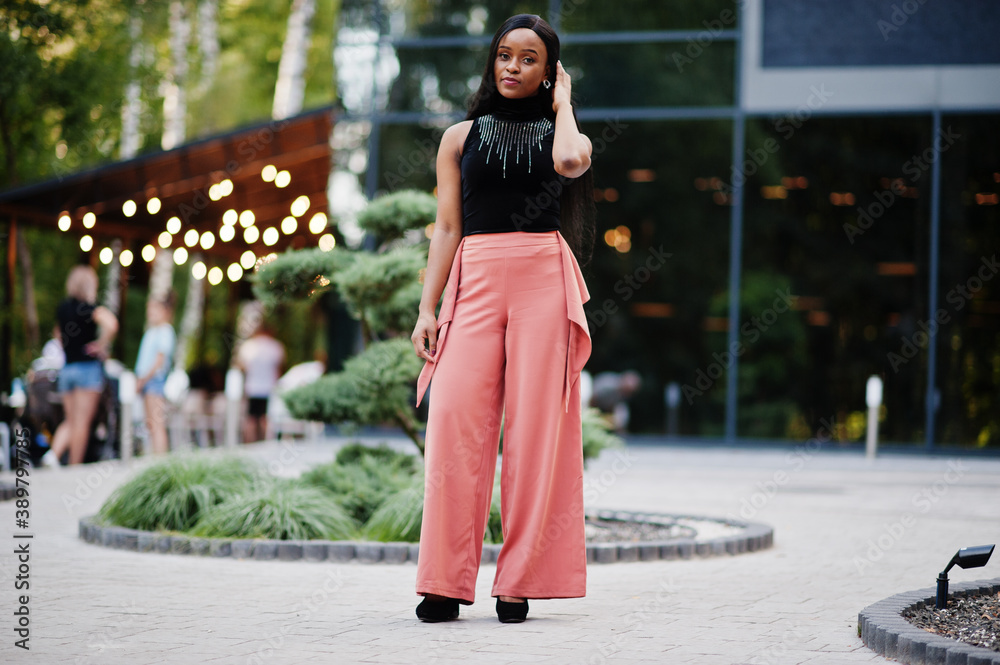 Fashionable african american woman in peach pants and black blouse pose outdoor.
