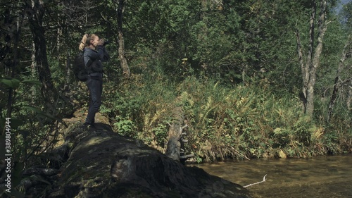 Traveler photographing scenic view in forest river. Wood bridge fallen tree. One caucasian woman shooting nice magic look. Girl take photo video on camera. Photographer travel with backpack. Outdoor.