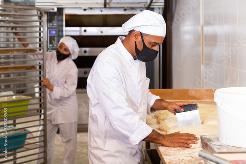 Worker of bakery in protective mask preparing fresh baked goods for sale on counter