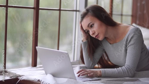 A young woman lies on the bed, looks at the laptop screen