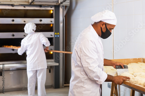 Worker of bakery in protective mask preparing fresh baked goods for sale on counter